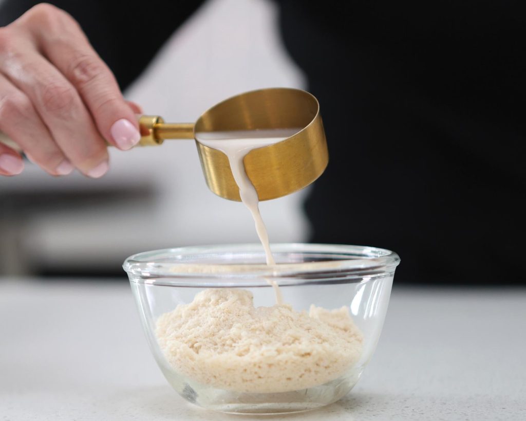 Front-facing photo of a hand pouring a cup of almond milk into a glass mixing bowl half filled with slice mix. 