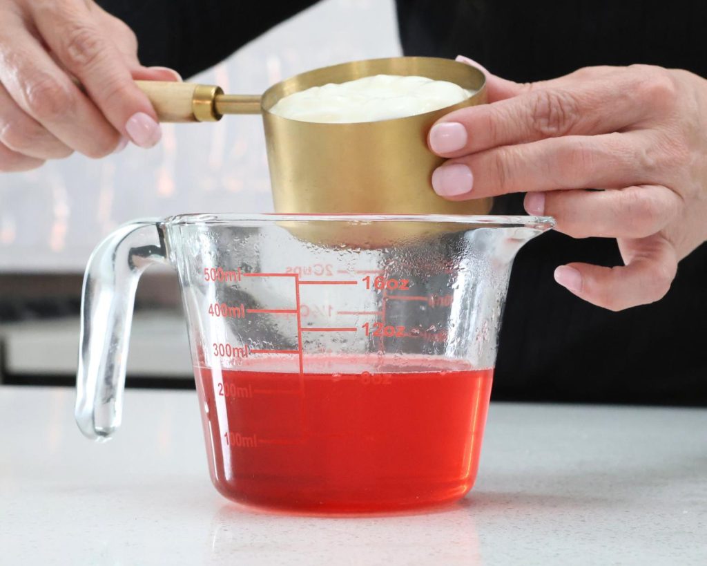 Front-facing photo of a hand pouring a cup of Greek yoghurt into a glass measuring jug half filled with protein jelly mix. 