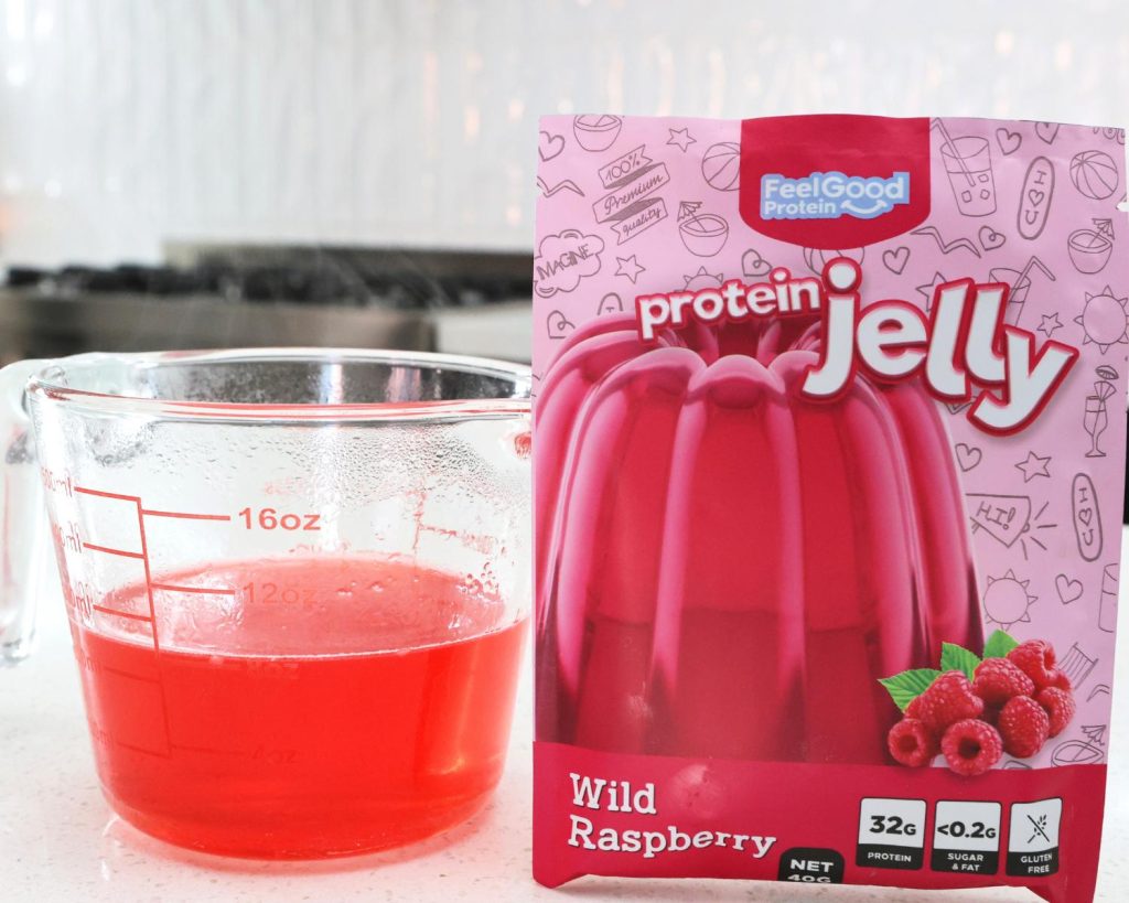 Front-facing photo of a glass measuring jug half filled with protein jelly mix next to a red sachet of Protein Jelly by Feel Good Protein. 