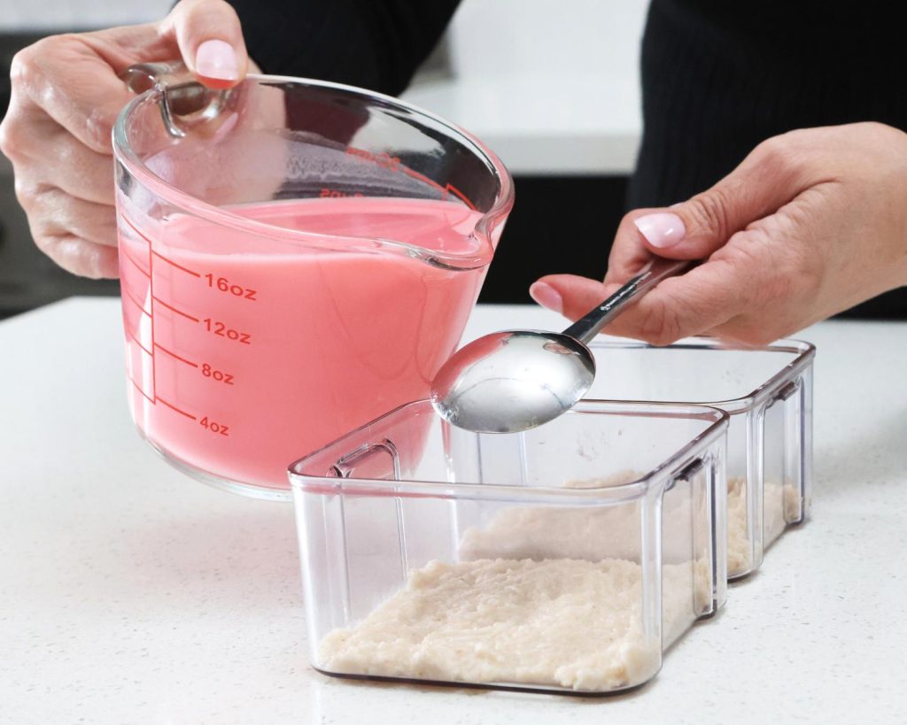 Front-facing photo of a hand pouring a glass measuring jug filled with pink cheesecake mixture over a spoon being held above two containers. 
