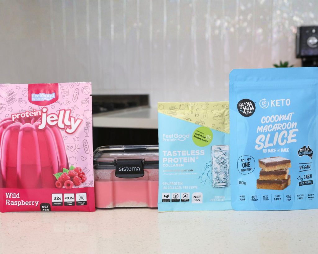 Front-facing photo of a red sachet of Protein Jelly by Feel Good Protein, next to a container of High Protein Raspberry Cheesecake Slice, next to a sachet of Tasteless Protein Collagen by Feel Good Protein, with a sachet of Coconut Macaroon Slice by Get Ya Yum On the right with a softly blurred modern kitchen in the background. 