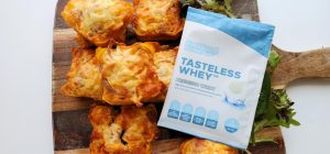 Birds eye view photo of a sachet of Tasteless Whey by Feel Good Protein in the centre of a wooden board holding 7 Bacon & Onion Protein Bites with a softly blurred modern kitchen in the background.