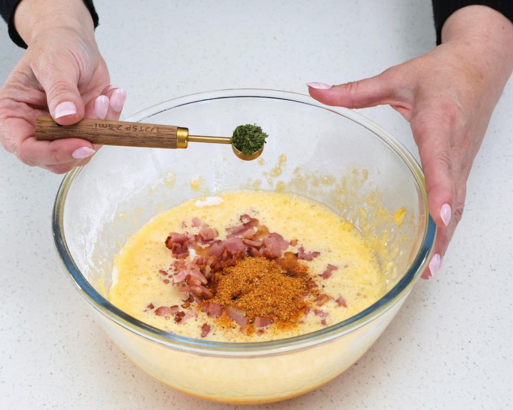 Front-facing photo of two hands holding a teaspoon of parsley over a glass mixing bowl filled with bacon and egg mix. 