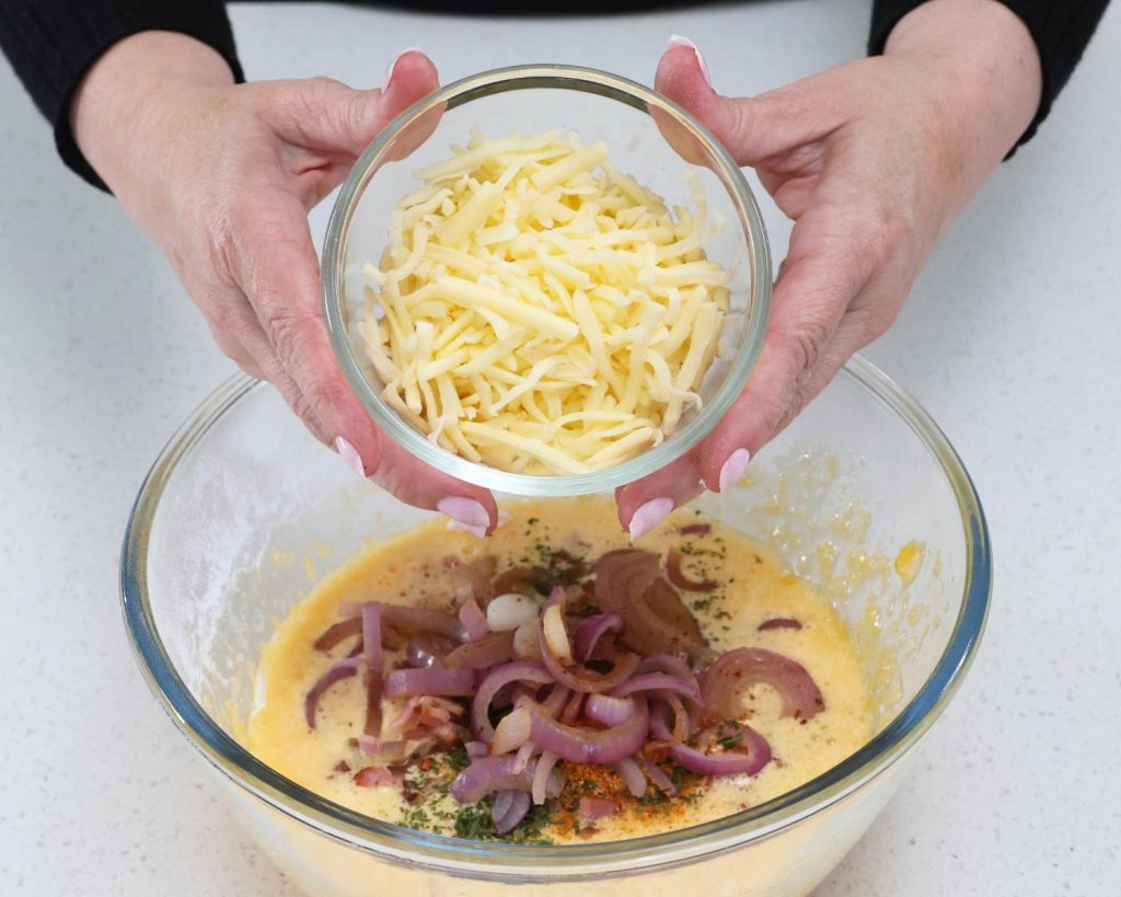 Birds eye view photo of two hands holding a small glass bowl of shredded cheese over a glass mixing bowl filled the Bacon & Onion Protein Bites mixture with a softly blurred modern kitchen in the background. 