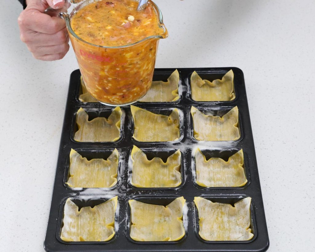 Birds eye view photo of a hand holding a glass measuring jug filled the Bacon & Onion Protein Bites mixture over a muffin tray filled with wonton wrappers. 