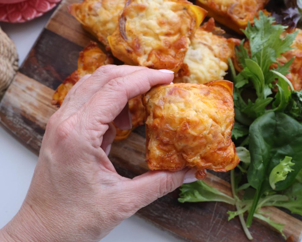 Birds eye view photo of a hand holding a Bacon & Onion Protein Bite mixture over the centre of a wooden board topped with Bacon & Onion Protein Bites with a softly blurred modern kitchen in the background. 