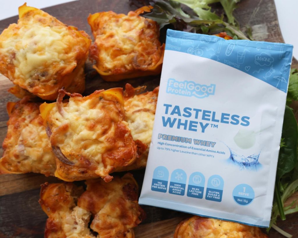 Birds eye view photo of a sachet of Tasteless Whey by Feel Good Protein in the centre of a wooden board holding 7 Bacon & Onion Protein Bites with a softly blurred modern kitchen in the background. 