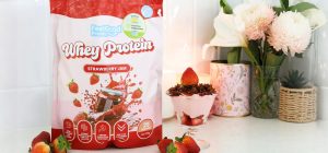 Front on photo of a red and pink back of Whey Protein by Feel Good Protein (Strawberry Jam) on the left of a Strawberry Cheesecake Protein Bowl placed on a kitchen bench.