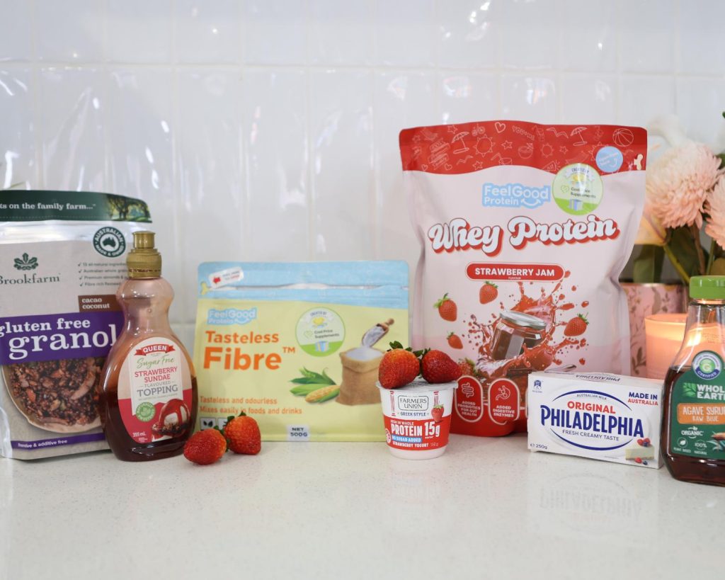 Front-facing photo showing all the ingredients for a Strawberry Cheesecake Protein Bowl recipe side by side on a benchtop, with a softly blurred modern kitchen in the background. 