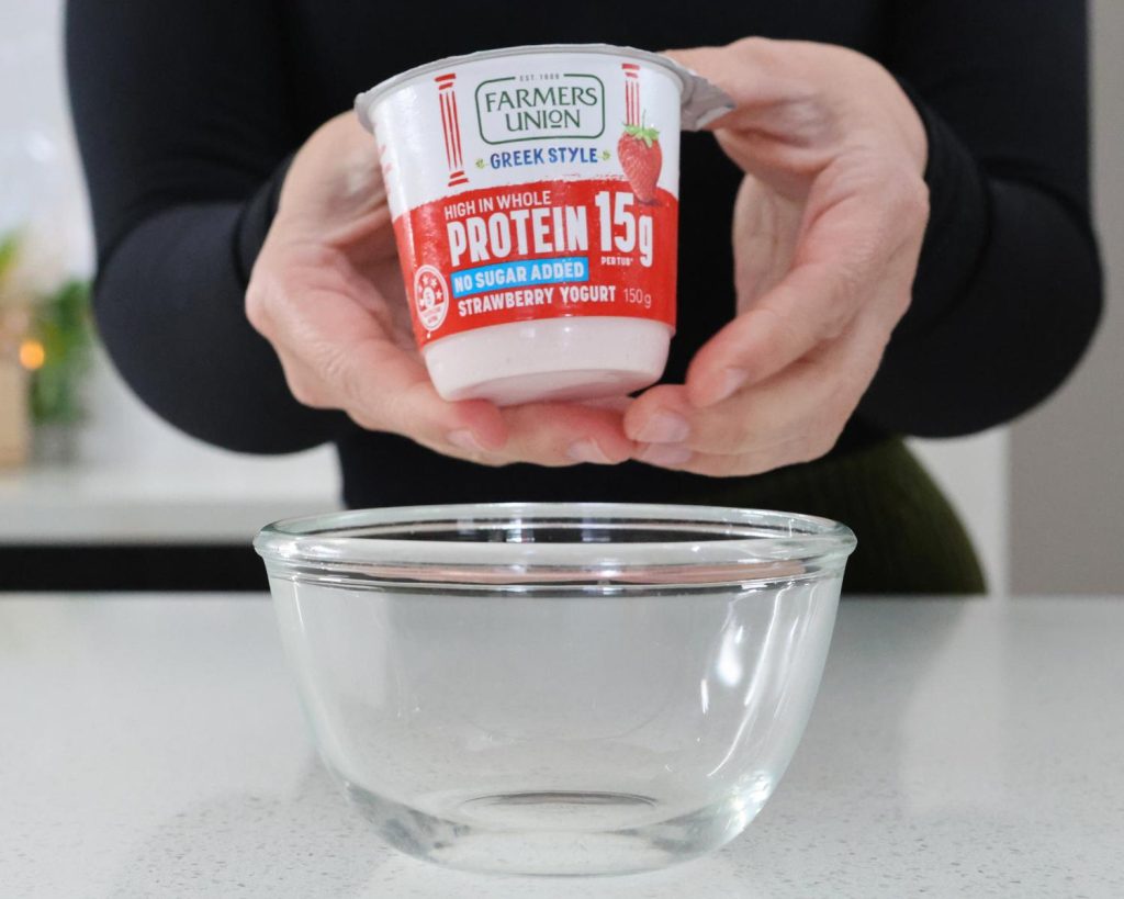Front on photo of two hands holding a 150g pink and white tub of strawberry yoghurt over a small glass mixing bowl. 