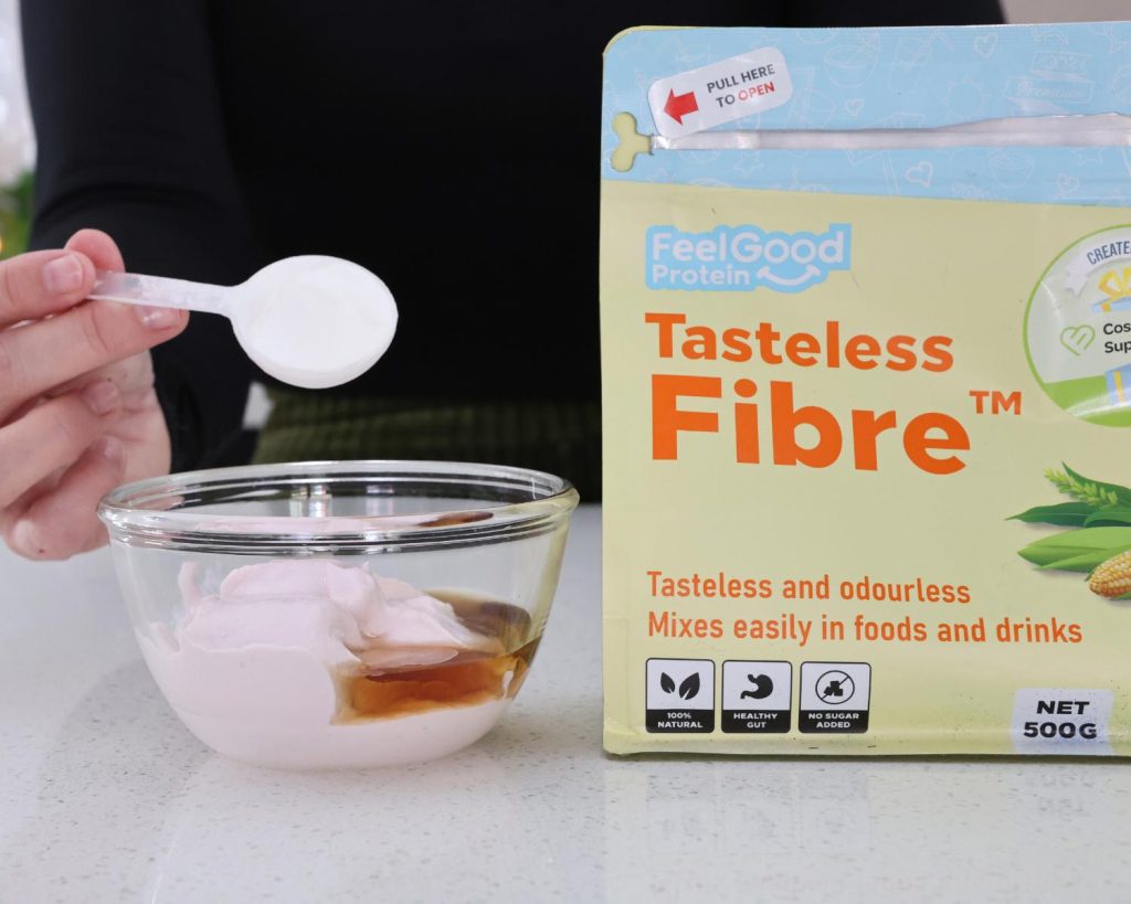 Front on photo of a hand holding a scoop over a glass mixing bowl half filled with strawberry yoghurt with a yellow and blue bag of Tasteless Fibre by Feel Good Protein on the right hand side. 