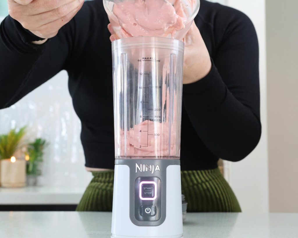 Front on photo of a hand holding a glass mixing bowl half filled with strawberry yoghurt over a Ninja blender. 