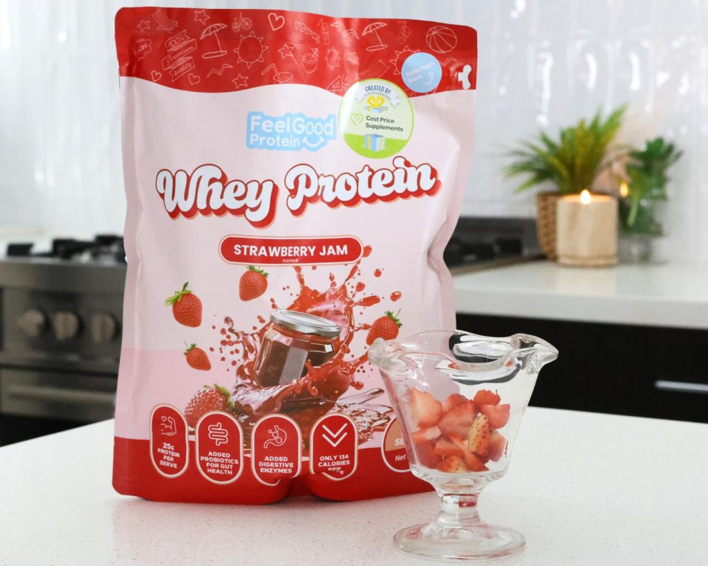 POV photo of a red and pink back of Whey Protein by Feel Good Protein (Strawberry Jam) behind a Strawberry Cheesecake Protein Bowl placed on a kitchen bench. 