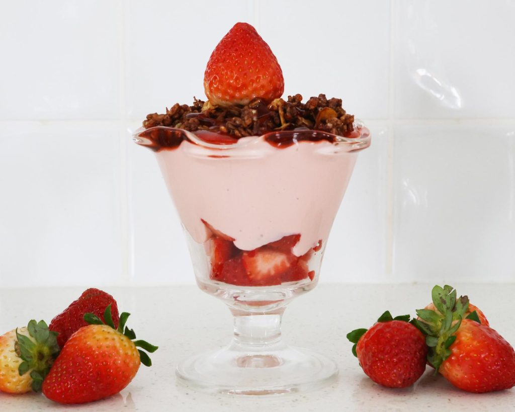 Photo of a Strawberry Cheesecake Protein Bowl placed on a kitchen bench with 3 strawberries lying on both sides of the bowl. 