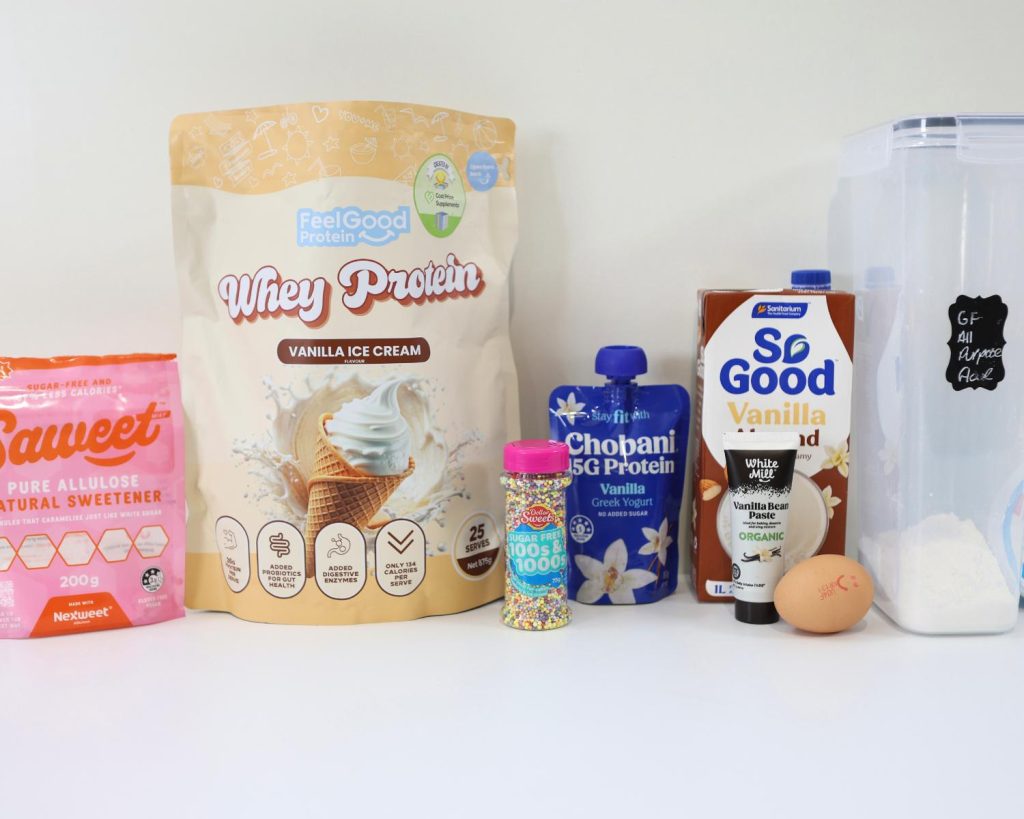 Front-facing photo showing all the ingredients for a High Protein Viral Dot Cake recipe side by side on a benchtop, with a softly blurred modern kitchen in the background. 