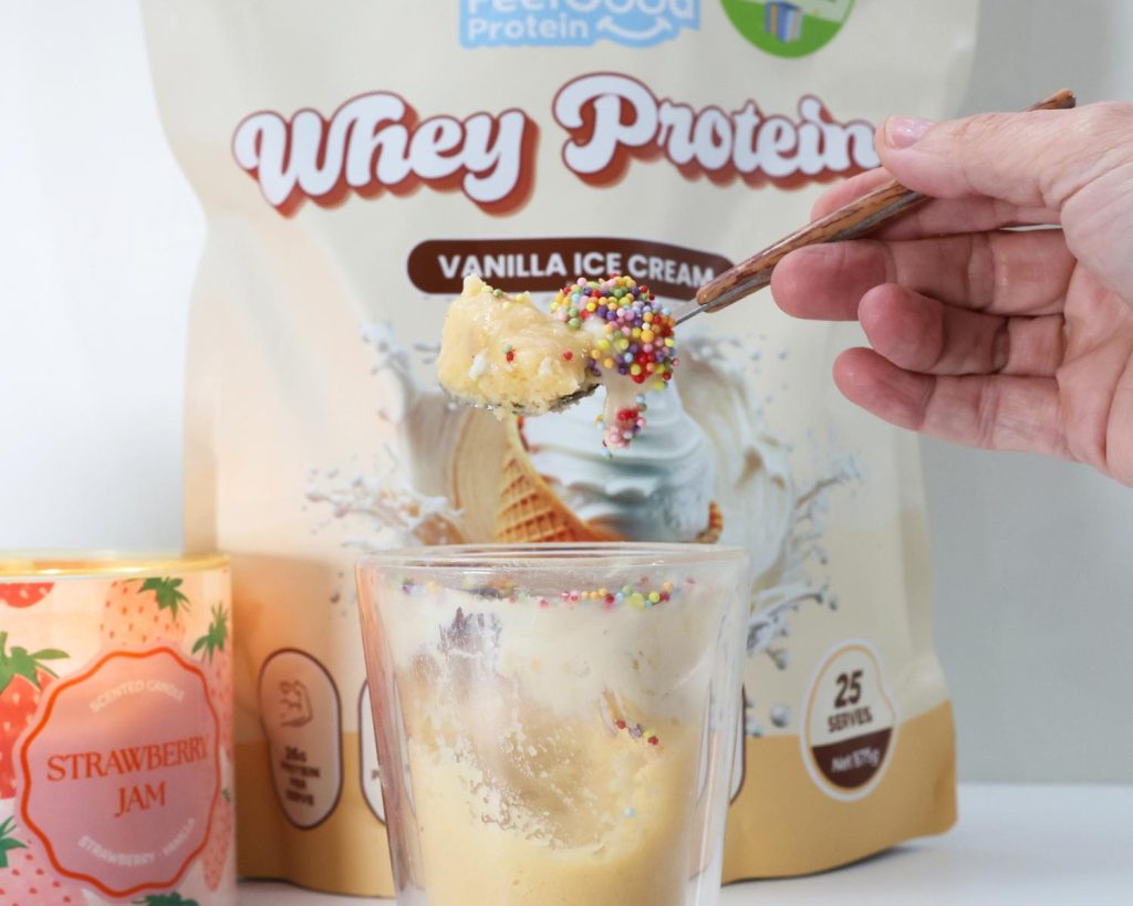 Front on photo of a yellow packet of Whey Protein by Feel Good Protein (Vanilla Ice Cream) behind a glass bowl of High Protein Viral Dot Cake with a hand holding a scoop of the cake over the rim. 