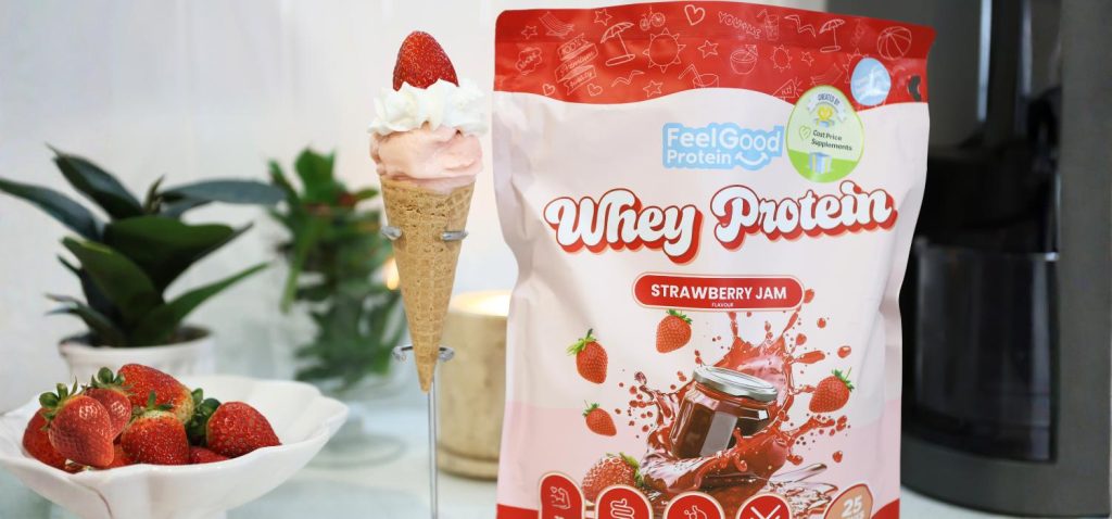 Front on photo of a red and pink packet of Whey Protein by Feel Good Protein (Strawberry Jam) on the right side of waffle cone of Ekka Strawberry Sundae placed on a kitchen bench.