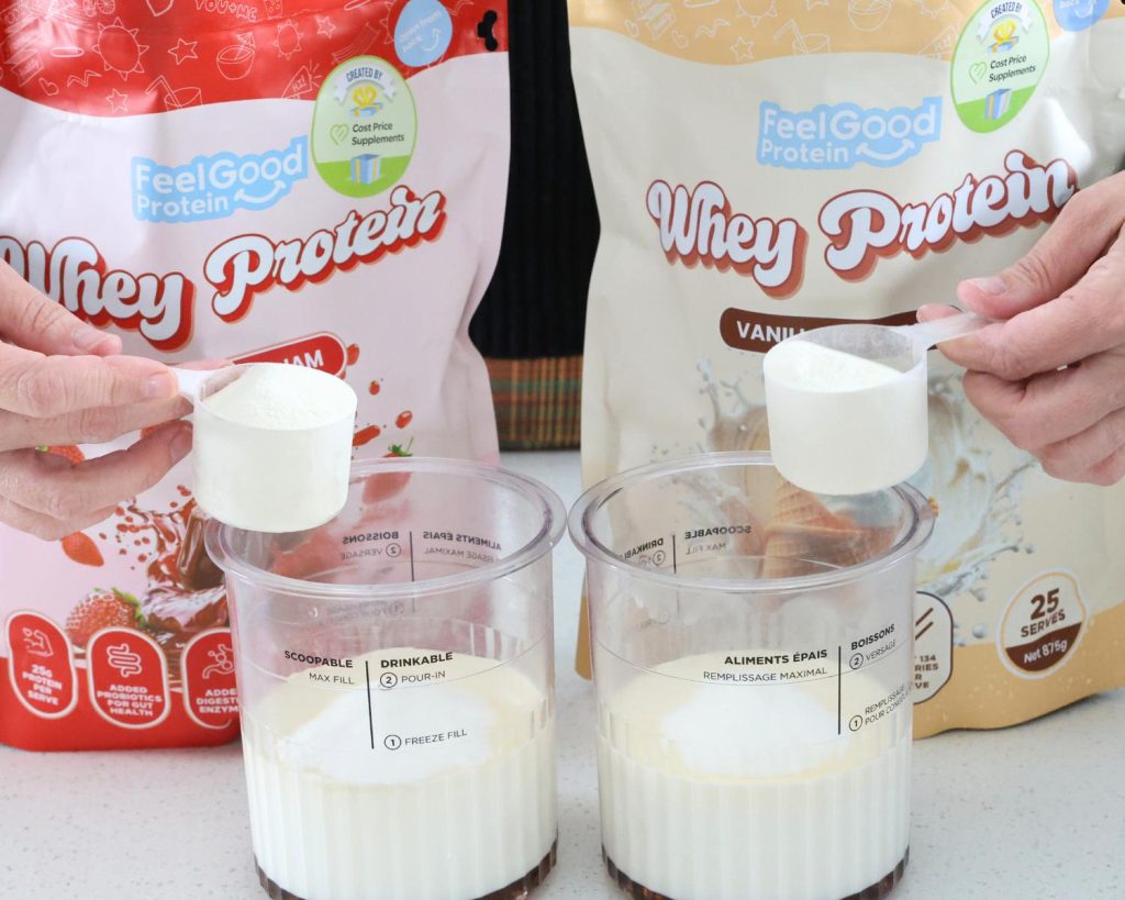 Front-facing photo of two hands holding two scoops of whey protein over two Ninja CREAMi tubs, in front of a red and pink packet of Whey Protein by Feel Good Protein (Strawberry Jam) and a yellow packet of Whey Protein by Feel Good Protein (Vanilla Ice Cream). 