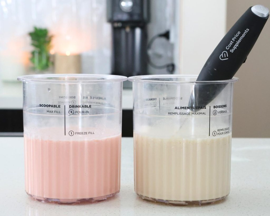 Front-facing photo of two Ninja CREAMi tubs with pink and white ice cream mixture, with a softly blurred modern kitchen in the background. 