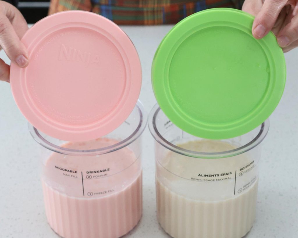 Front-facing photo of two hands covering two Ninja CREAMi tubs filled with pink and white ice cream mixtures, with pink and green lids. 