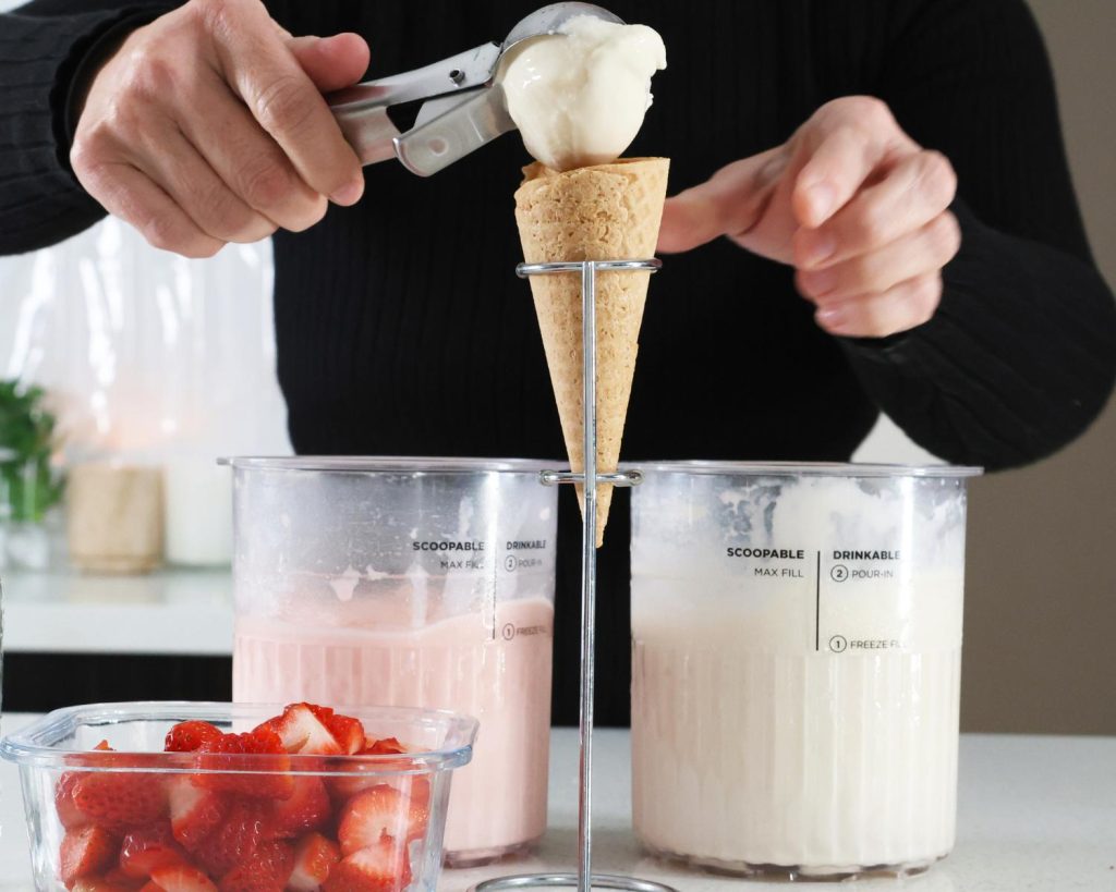 Front-facing photo of a hand scooping ice cream from two Ninja CREAMi tubs filled with pink and white ice cream mixtures, into a waffle cone