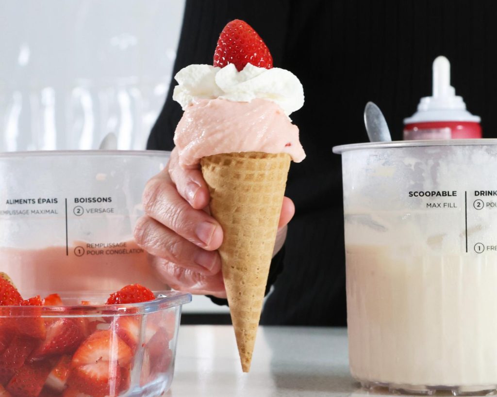 Front-facing photo of a hand holding an Ekka Strawberry Sundae in between two Ninja CREAMi tubs filled with pink and white ice cream mixtures. 