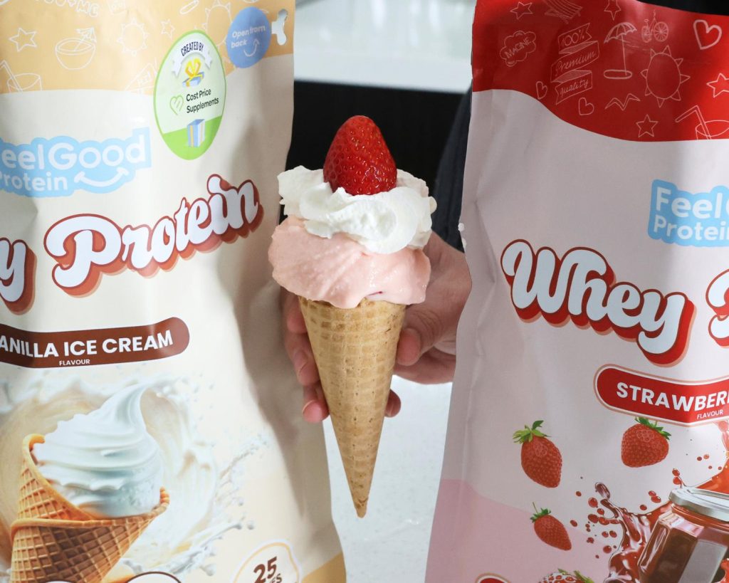 Front-facing photo of a hand holding an Ekka Strawberry Sundae in between a red and pink packet of Whey Protein by Feel Good Protein (Strawberry Jam) and a yellow packet of Whey Protein by Feel Good Protein (Vanilla Ice Cream). 